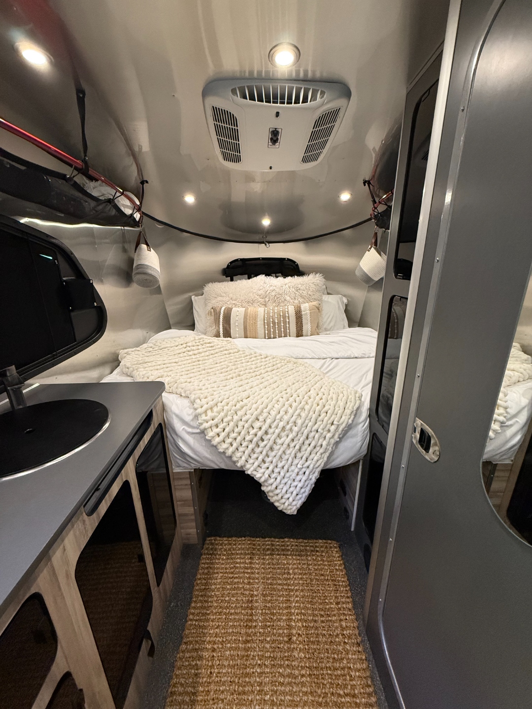 2023 Airstream Basecamp 20X rear king bed dressed with the optional Glamping Bedding Package — chunky knit throw, boho pillows, and jute rug